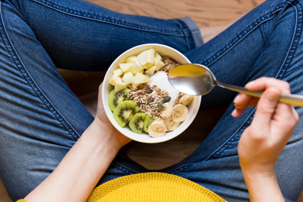 A 16 weeks pregnant woman sat cross legged in blue jeans holding a bowl of yoghurt, oats and fruit