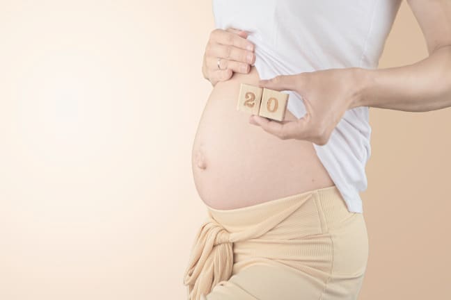 A pregnant woman in a white top and nude coloured bottoms holding 2 blocks with number 20 engraved