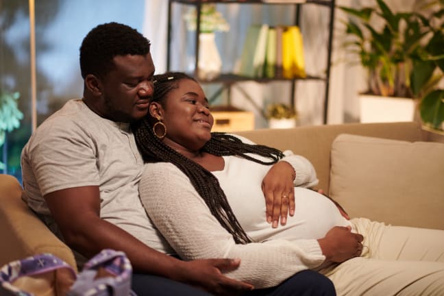 A black couple on the sofa, with the man wrapping his arms around his pregnant wife 