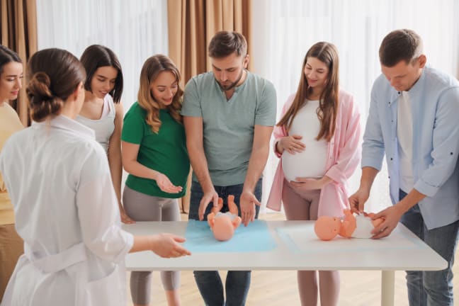 A group of 5 pregnant women and 2 men, attending a baby class learning how to change a nappy 