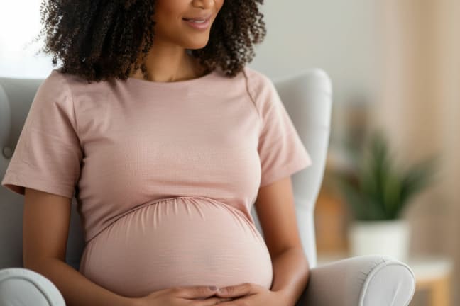 A pregnant woman in a pink coloured top sat in a grey armchair holding onto her belly