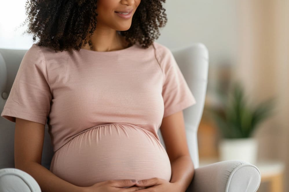 A woman in a pink coloured top who is 25 weeks pregnant sat in an armchair holding onto her belly.