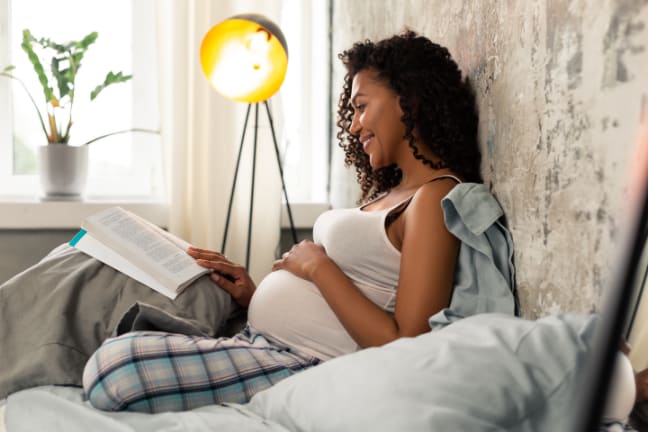 A woman who is 26 weeks pregnant sat upright in bed reading a book.