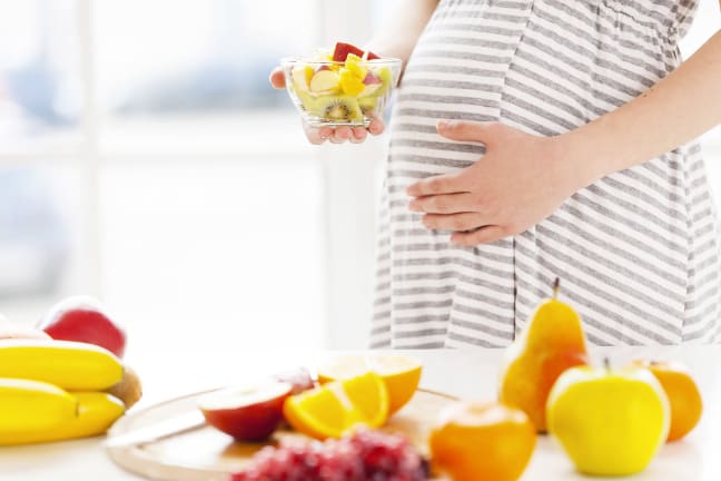 A 27 weeks pregnant woman in front of a white kitchen work top making a bowl of fruit