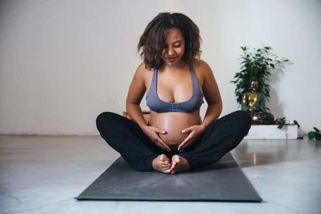 A pregnant woman in a grey sports bra and black bottoms, sat in a yoga pose holding onto her belly