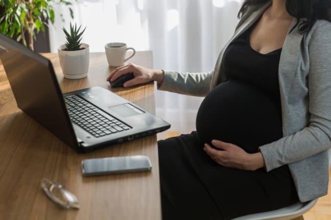 A woman who is 29 weeks pregnant sat at a desk looking at her laptop holding onto her belly 