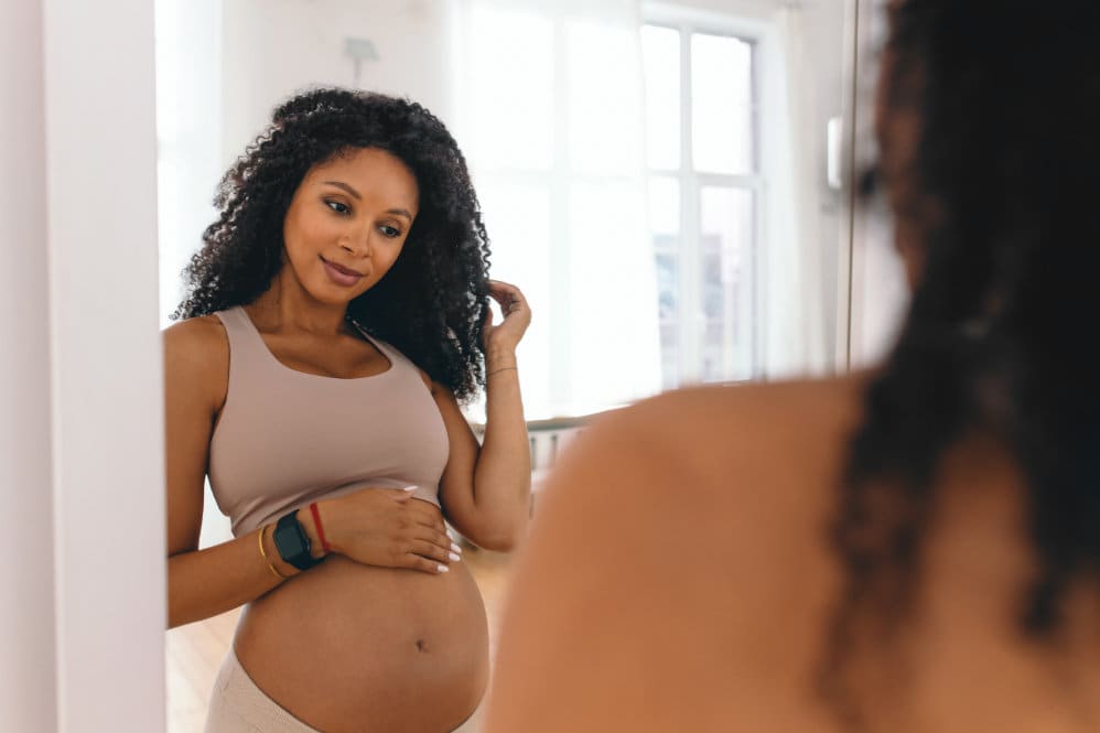 A pregnant woman in a nude sports bra, looking at a reflection of her pregnant belly in the mirror