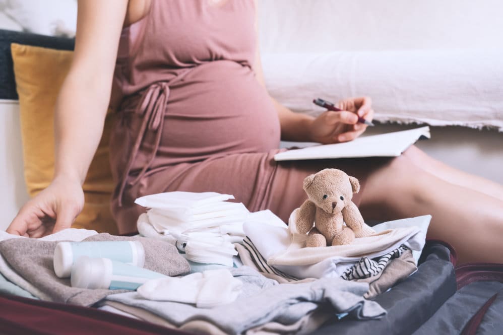 A pregnant woman wearing a pink dress, preparing her hospital bag whilst writing down a checklist