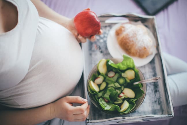 A pregnant woman in a white top sat on the bed with a tray of healthy food, also holding an apple