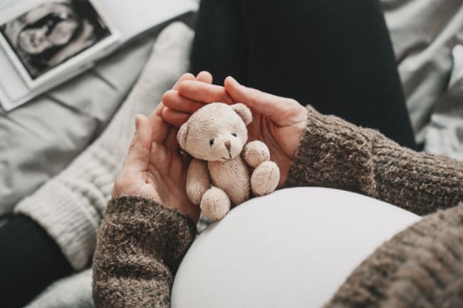 A 36 weeks pregnant woman in a white top looking down at her baby scan, holding onto a teddy bear