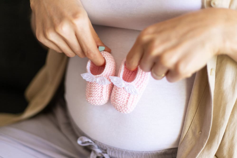 A 39 weeks pregnant woman in a white top and linen shirt holding a pair of pink, knitted booties