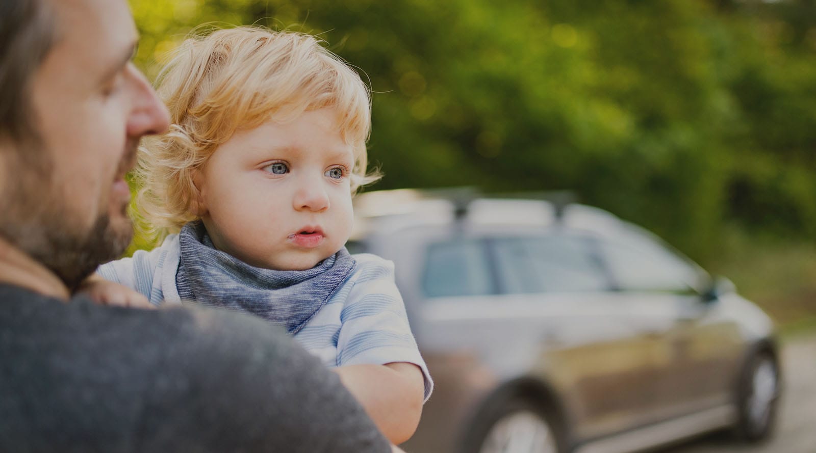 A toddler in his father's arms gazing into the distance, with a car in the background.