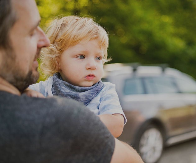 A toddler in his father's arms gazing into the distance, with a car in the background.
